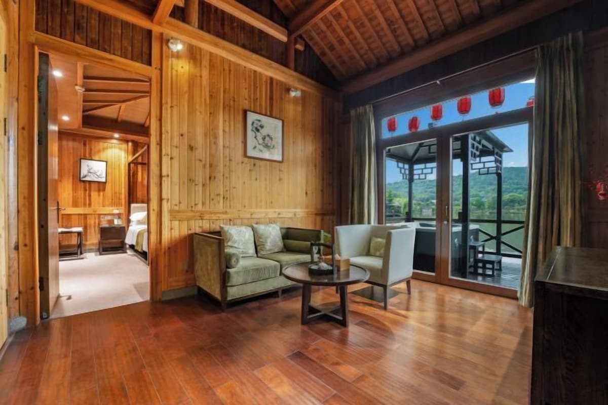 A wooden interior with a sofa, armchair, and table. There is a view through a glass door to the outside.