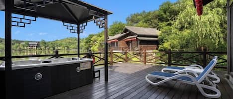 A wooden deck with a hot tub and lounge chairs, overlooking a traditional building and lush greenery.