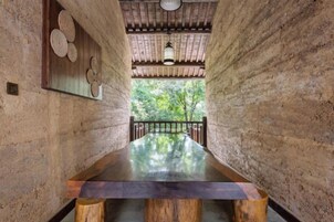 Una larga mesa de madera en una habitación rústica con paredes de ladrillo visto y vistas al exterior con vegetación.