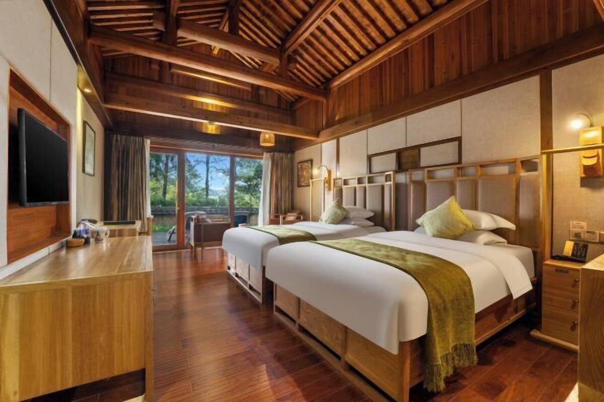 A hotel room with two beds, wooden flooring, a TV, and a balcony with a view of trees.