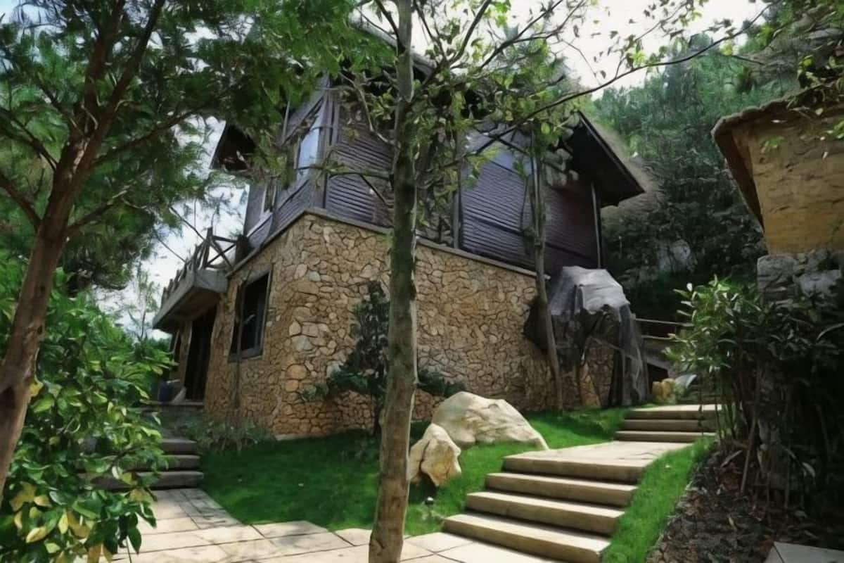 A stone house with a staircase leading to a wooden deck, surrounded by greenery.