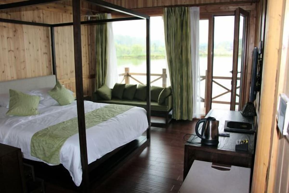 A four-poster bed with green bedding, a wooden desk with a kettle, and a view of a wooden deck and greenery.