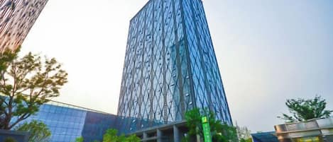 A tall, modern building with a unique facade, surrounded by greenery and a clear sky.