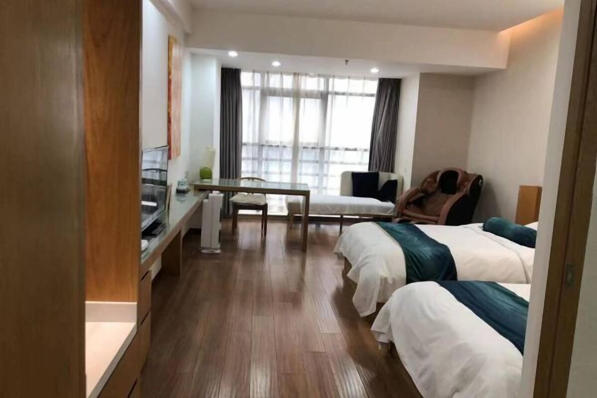 A hotel room with a wooden floor, a desk with a chair, two beds with white and teal bedding, and a large window with curtains.