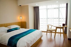 Una habitación de hotel con cama, una mesa, una silla, un ventanal con cortinas y una lámpara fijada a la pared.