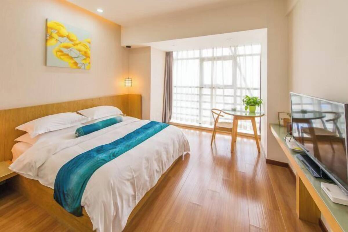 A hotel room with a bed, a desk with a TV, a window with curtains, and a painting on the wall.