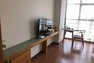 Una habitación con piso de madera, un televisor sobre un soporte de madera, una pequeña mesa con sillas y un ventanal grande.