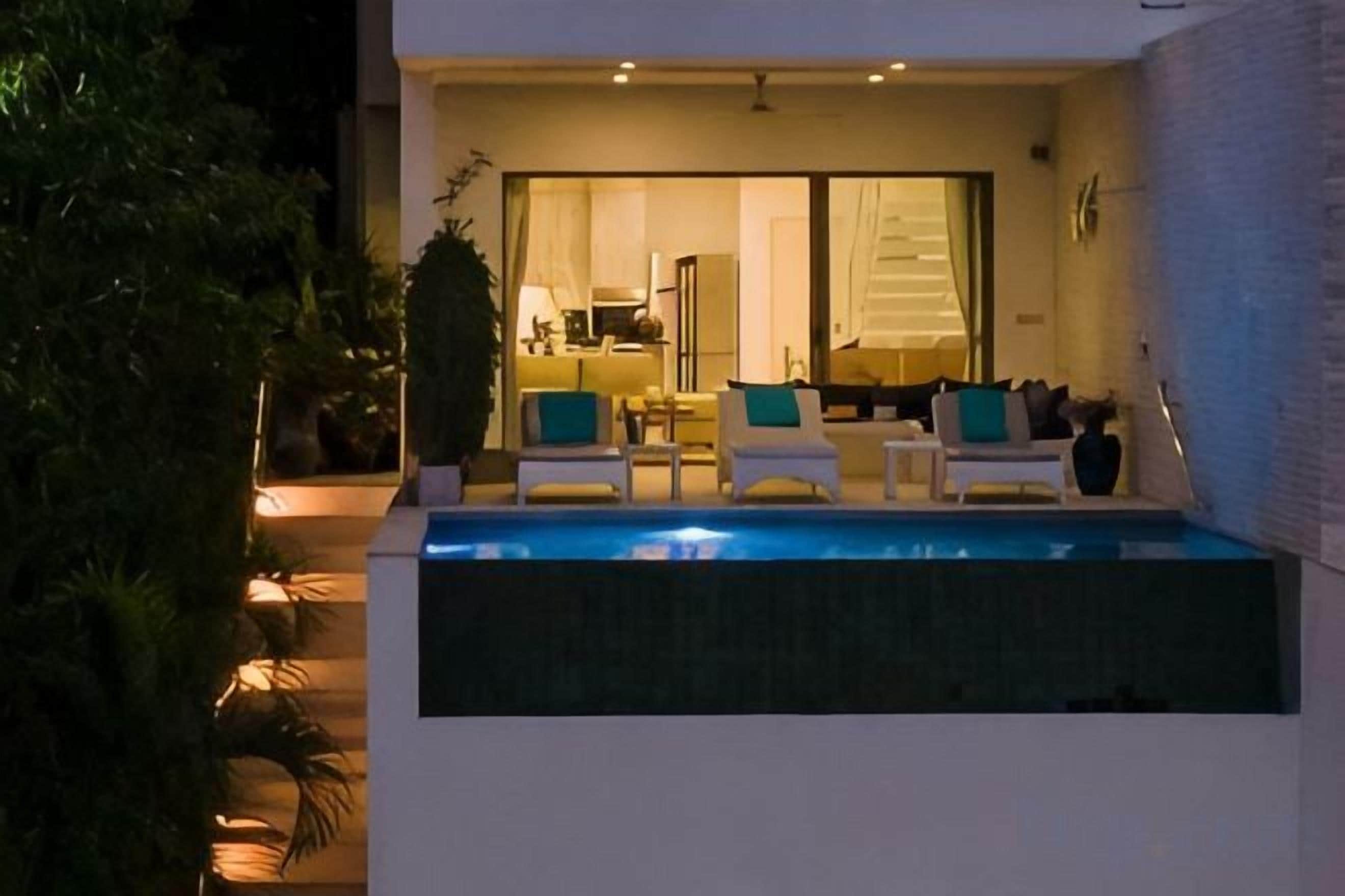 A modern outdoor pool area with lounge chairs and a dining area visible through a glass door.