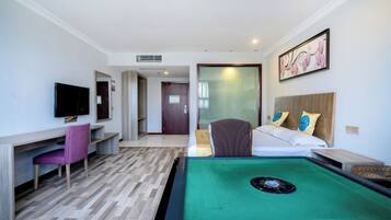 A hotel room with a pool table, a TV, a desk with a chair, a bed with pillows, and a door leading to another room.