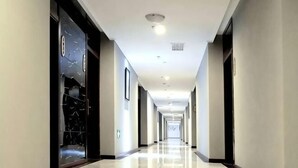 Un lungo e stretto corridoio d'albergo con pavimento lucido, porte in vetro a cornice scura e illuminazione a incasso.