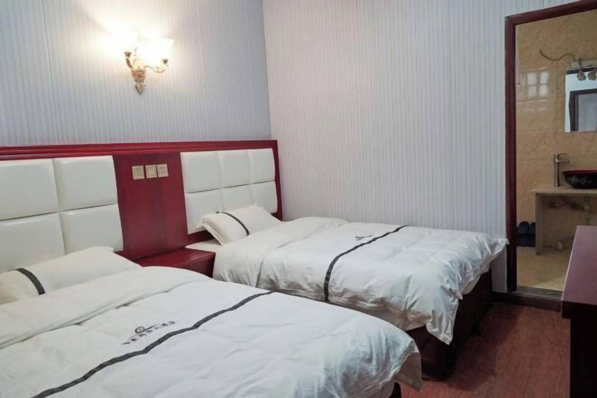 A hotel room with two beds, a red headboard, a wall-mounted light fixture, and a small bathroom area visible through an open door.