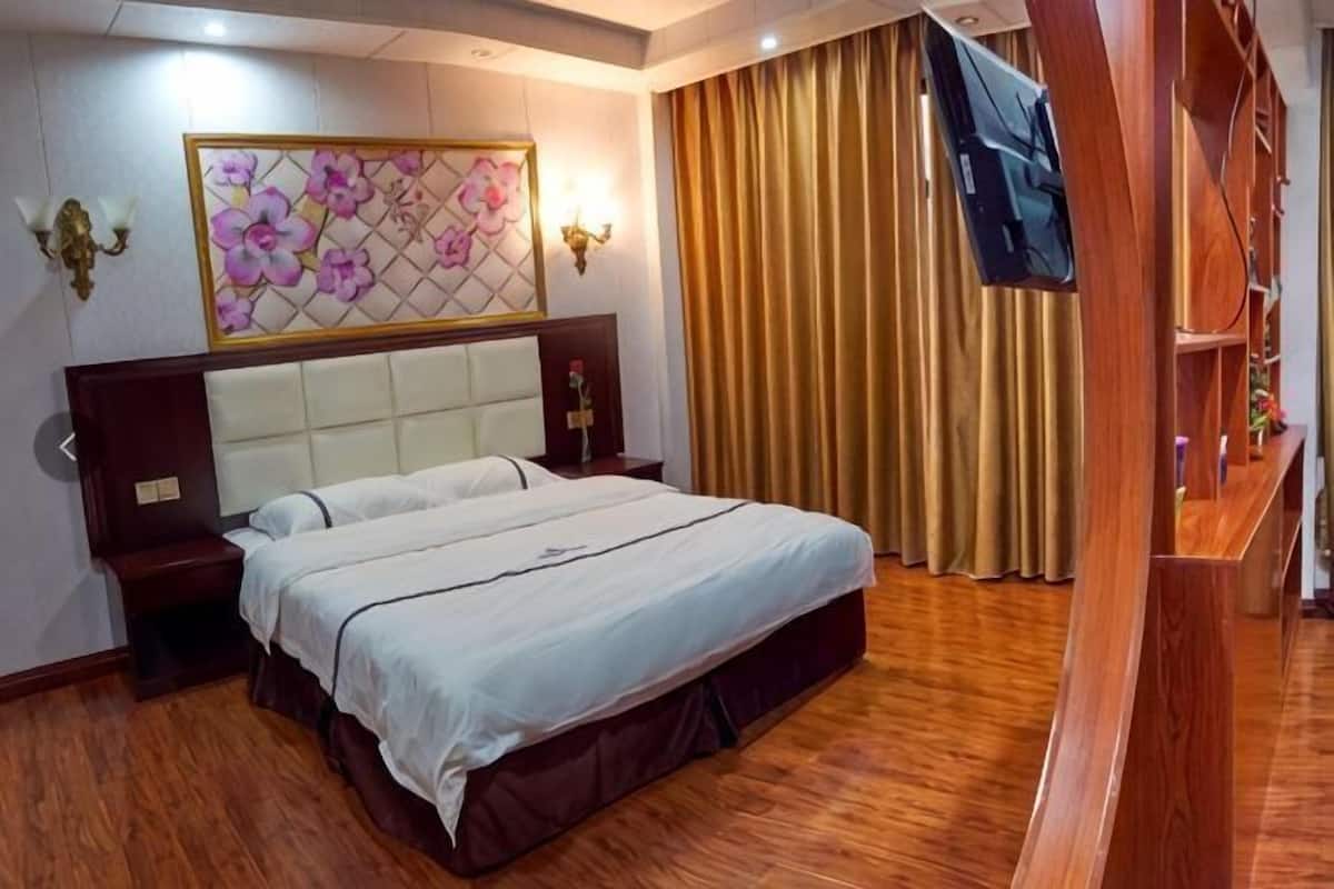 A hotel room with a large bed, wooden flooring, a headboard with a floral design, a television, and a window with curtains.