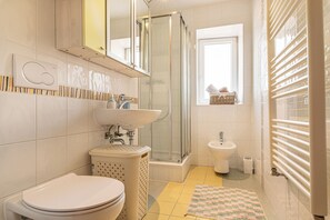 A bathroom with a toilet, sink, bidet, shower, and a window with a basket on the sill. - Studio Izola Near the old Town (Izola)