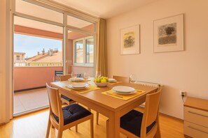 A dining area with a wooden table set for four, a bowl of fruit, and a view of buildings through large windows. - Studio Izola Near the old Town (Izola)