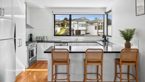 Een moderne keuken met een groot raam, witte kasten, een centraal eiland met barkrukken en uitzicht op woningen.