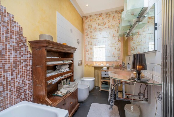 A bathroom with a bathtub, a wooden shelf with towels, a toilet, and a sink with a mirror.