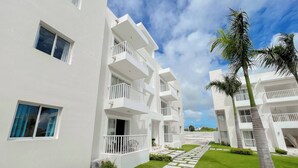 A modern white building with multiple balconies, a well-maintained lawn, and palm trees. - Goistay Punta Cana Sol, Playa Pool 15 Min Airport (Punta Cana)