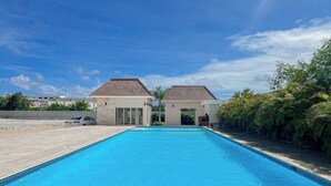 A rectangular swimming pool with clear blue water, surrounded by a concrete deck. There are two small buildings with tiled roofs and glass doors, one on each side of the pool. The area is landscaped with green shrubs and palm trees. - Goistay Punta Cana Sol, Playa Pool 15 Min Airport (Punta Cana)