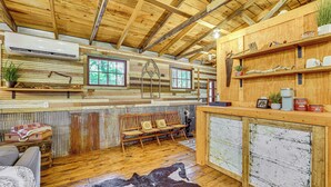 A rustic cabin interior with wooden beams, a wooden bench, a cowhide rug, and a rustic bar area with shelves. - Unique Eureka Springs Cabin: Deck & Forest Views! (Busch)