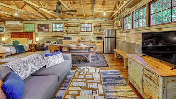 A spacious living area with a sofa, coffee table, and a dining table. There is a kitchen area with a refrigerator and a large window. - Unique Eureka Springs Cabin: Deck & Forest Views! (Busch)