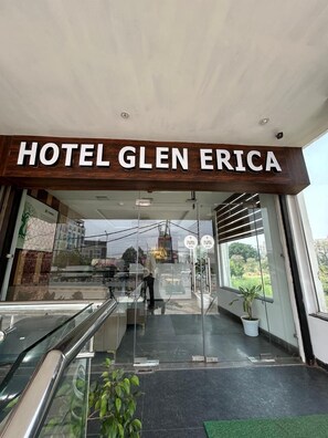 The entrance to Hotel Glen Erica with a clear view of the reception area, including a reception desk and a glass door. - Hotel Glen Erica (Dera Bassi)