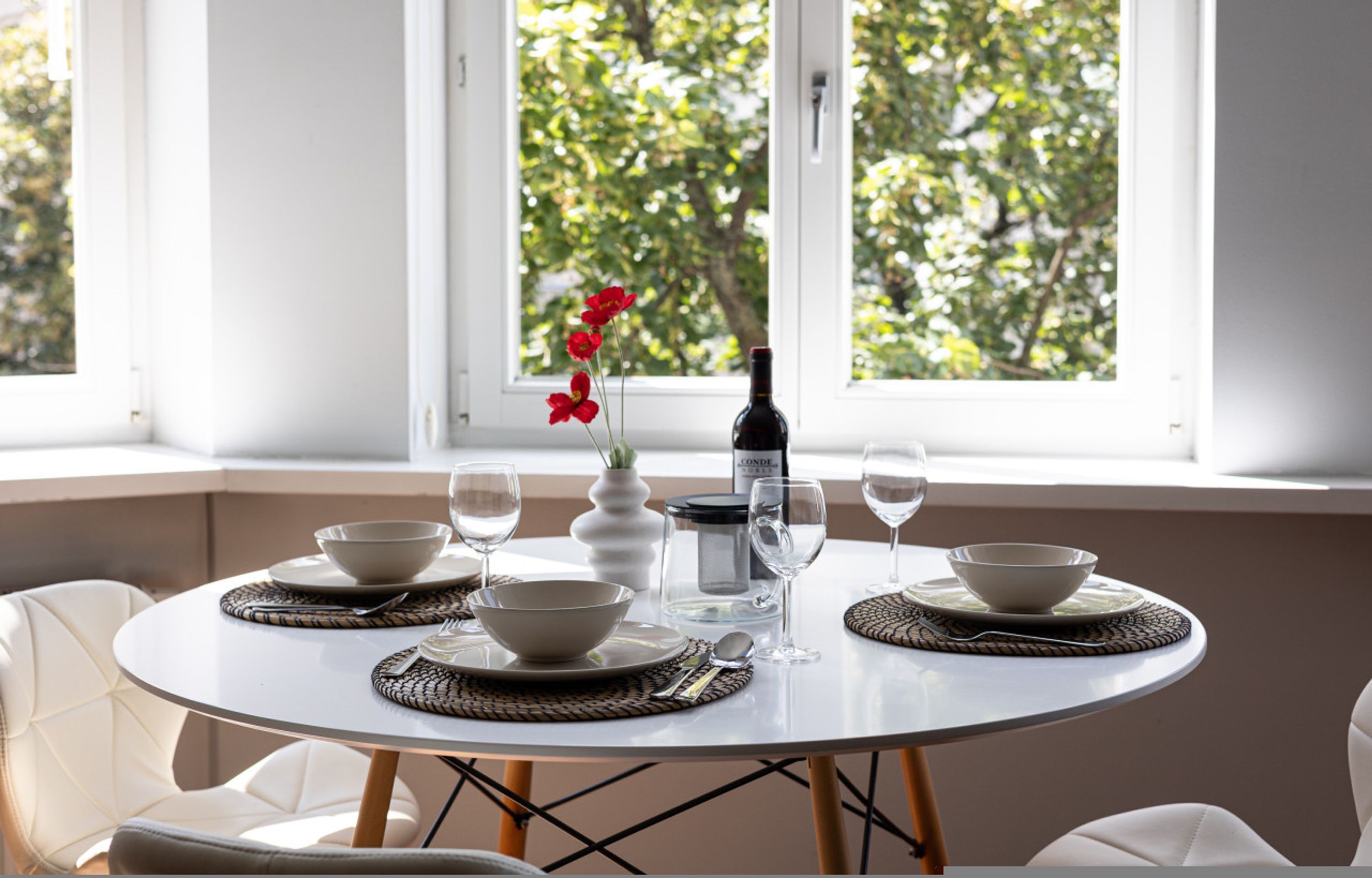 A round dining table with white chairs, a vase with red flowers, a bottle of wine, and several wine glasses.