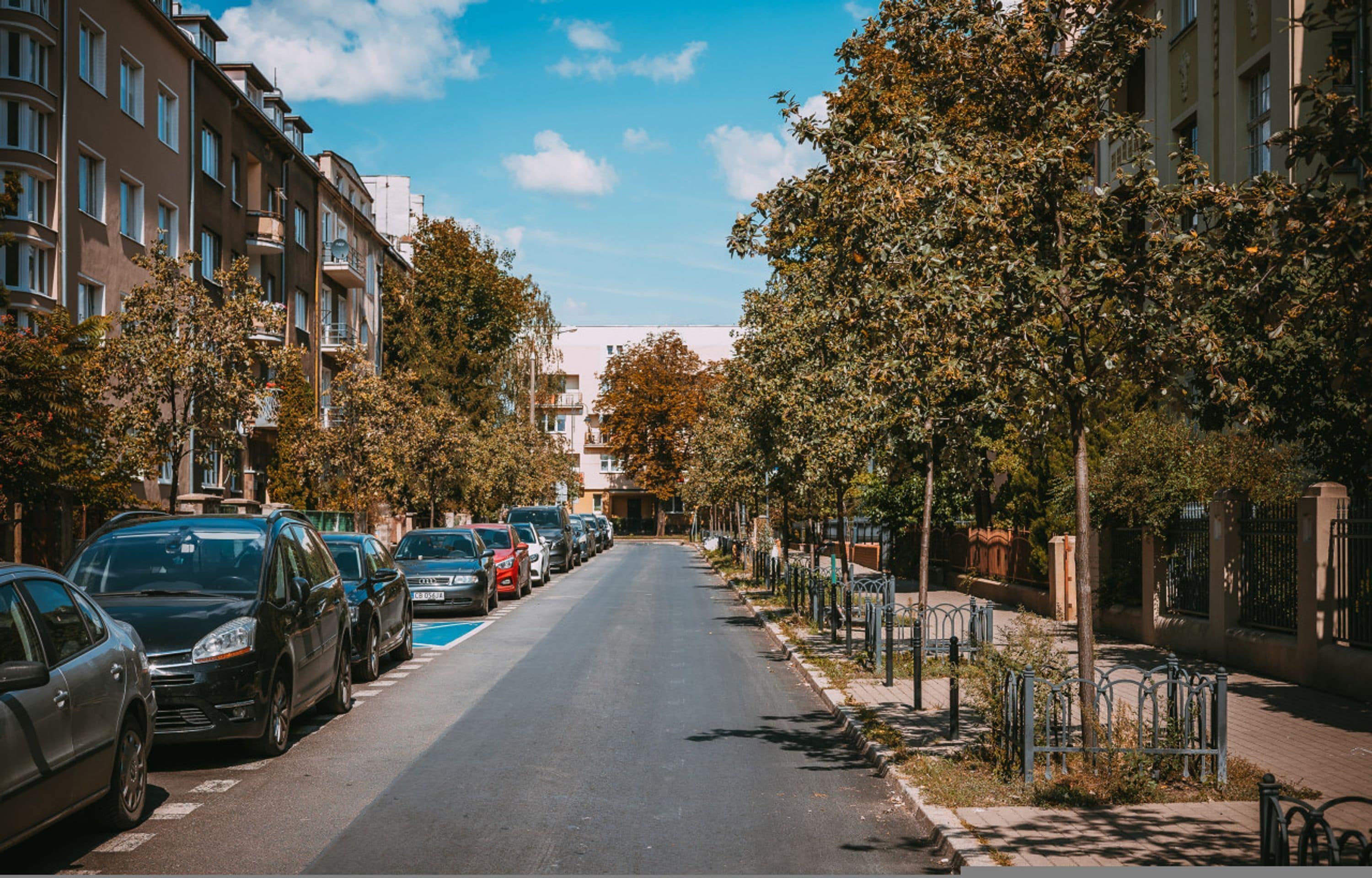 A street lined with parked cars and trees, leading to a building with multiple floors and balconies.
