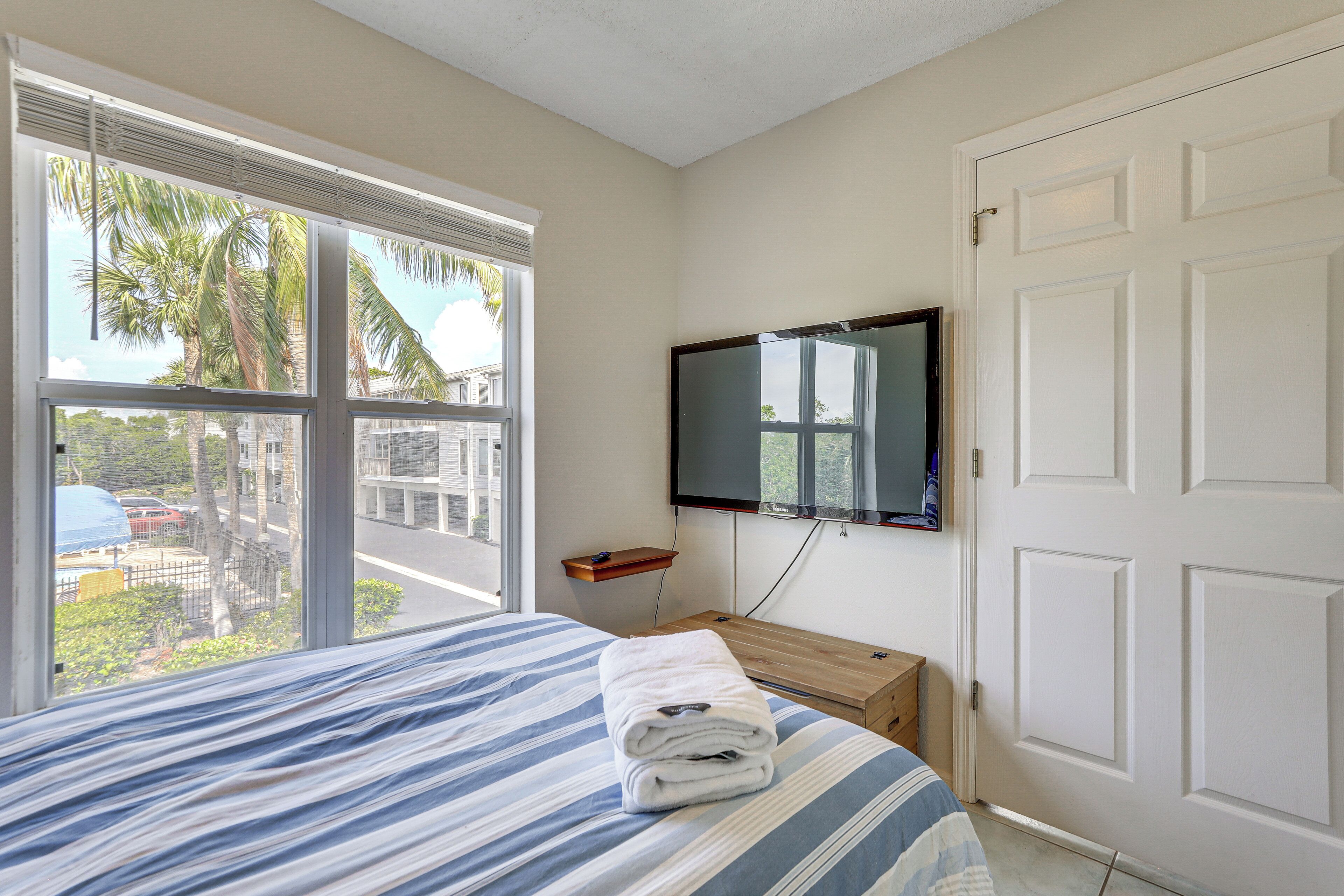 Un dormitorio con cama, televisor montado en la pared, ventana con persianas y vista a palmeras y una calle.