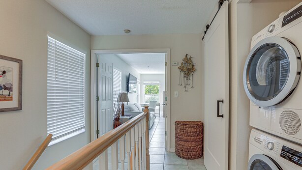 A laundry room with stacked washers and dryers, a wicker basket, and a hallway leading to a living area with a television and a sofa. - Holmes Beach Condo Near Anna Maria: Pool Access! (Holmes Beach)