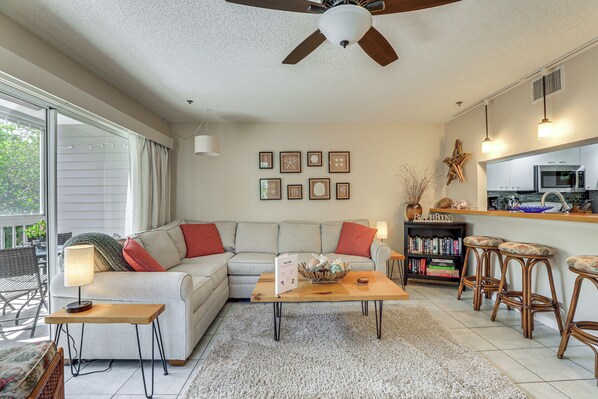 A living room with a sofa, coffee table, and a dining area with bar stools. There is a kitchen in the background with a microwave and a starfish decoration. - Holmes Beach Condo Near Anna Maria: Pool Access! (Holmes Beach)