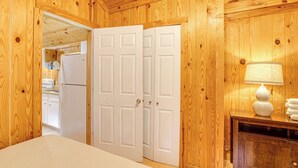Una habitación con paneles de madera, una cama, una puerta blanca y una lámpara sobre una mesa de madera.