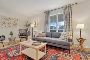 Una sala de estar con un sofá gris, un tapete rojo estampado, una mesita de centro de madera y una ventana con cortinas.