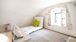 Ein Zimmer mit einem weißen Bett, einem grünen Kopfkissen, einem kleinen Fenster und einer weißen Wand.