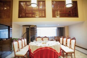 Un comedor con una mesa redonda dispuesta para una comida, rodeada de sillas, con una escalera y un televisor al fondo.