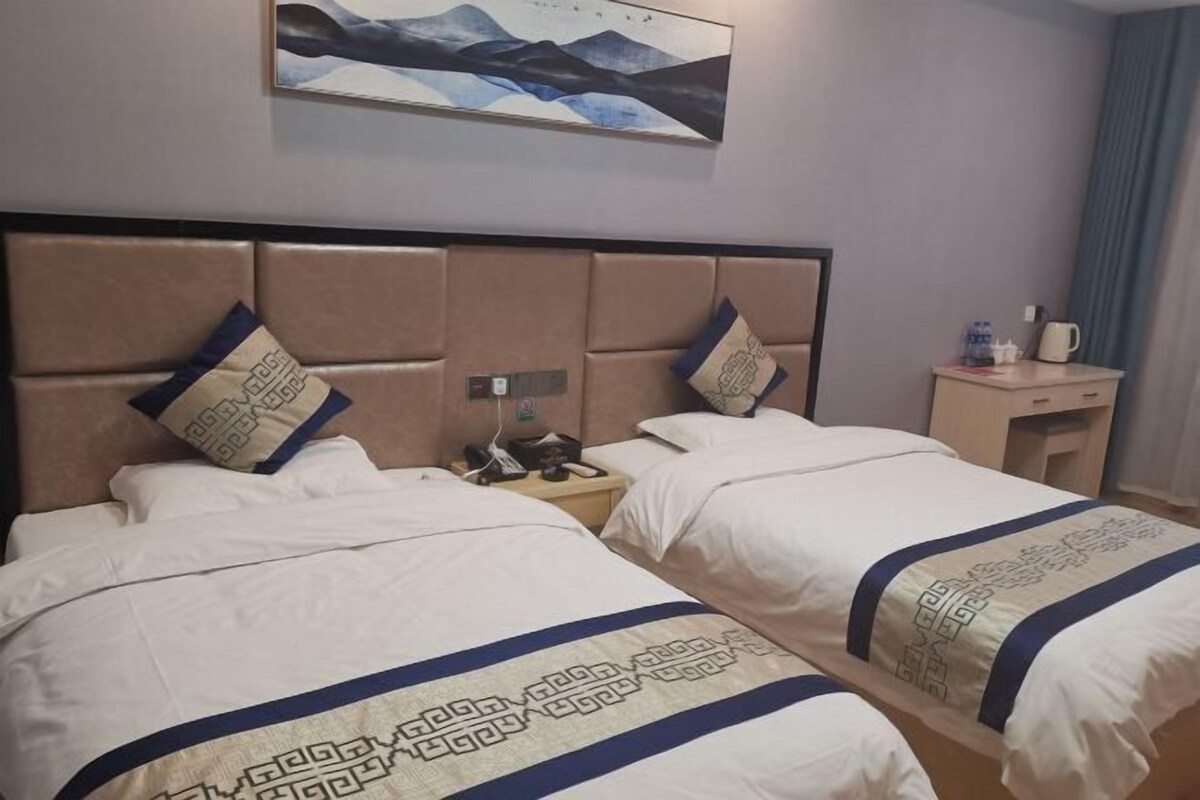 Habitación de hotel con dos camas, cada una con cabecera y almohadones decorativos. Hay una mesita con un teléfono y una taza, y un cuadro sobre las camas.