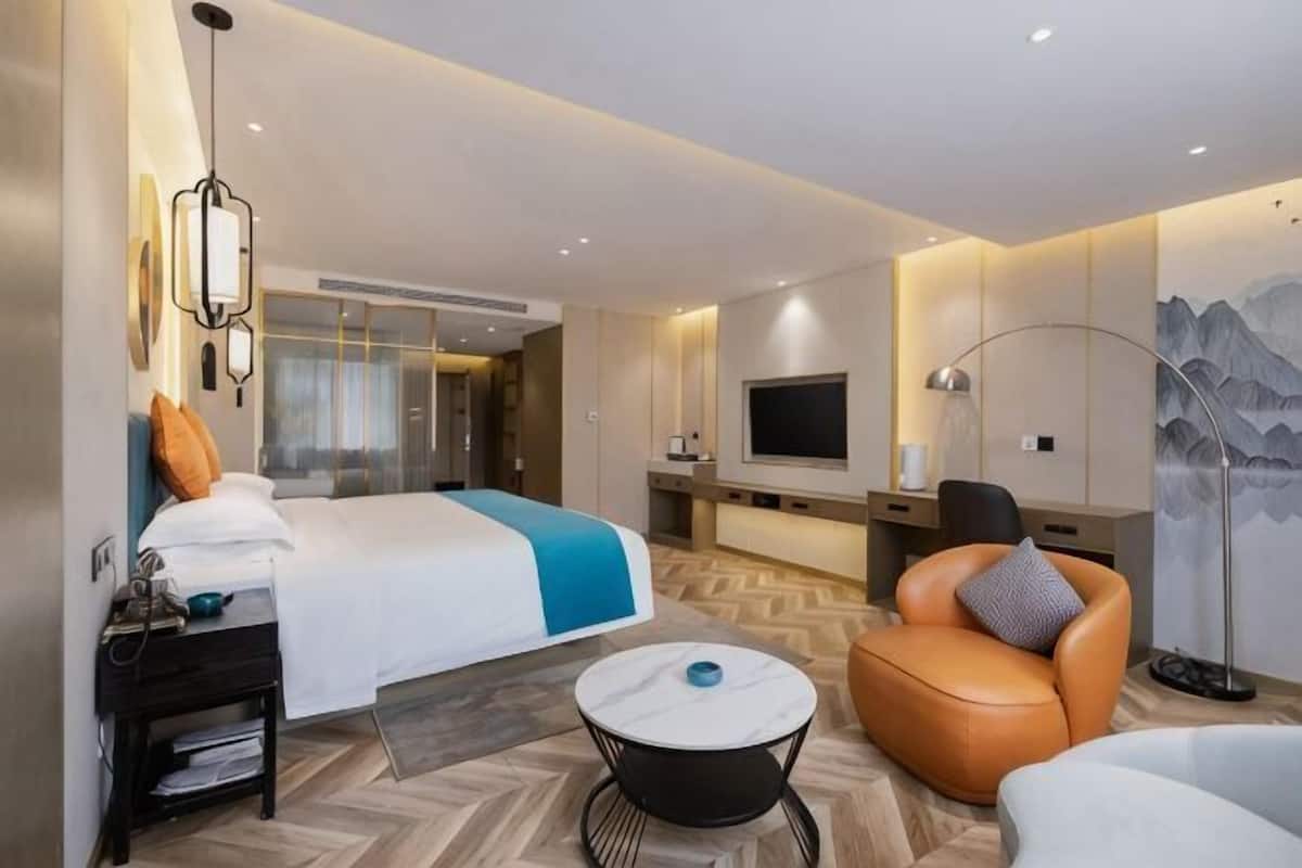 Una habitación de hotel con una cama, una zona de estar con una silla y una mesita, un televisor y una lámpara fijada a la pared.