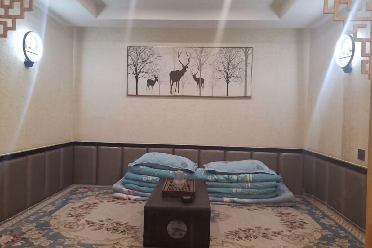 Habitación de esquina con alfombra estampada, cama con ropa de cama azul, mesita de noche con lámpara y un cuadro de ciervos.