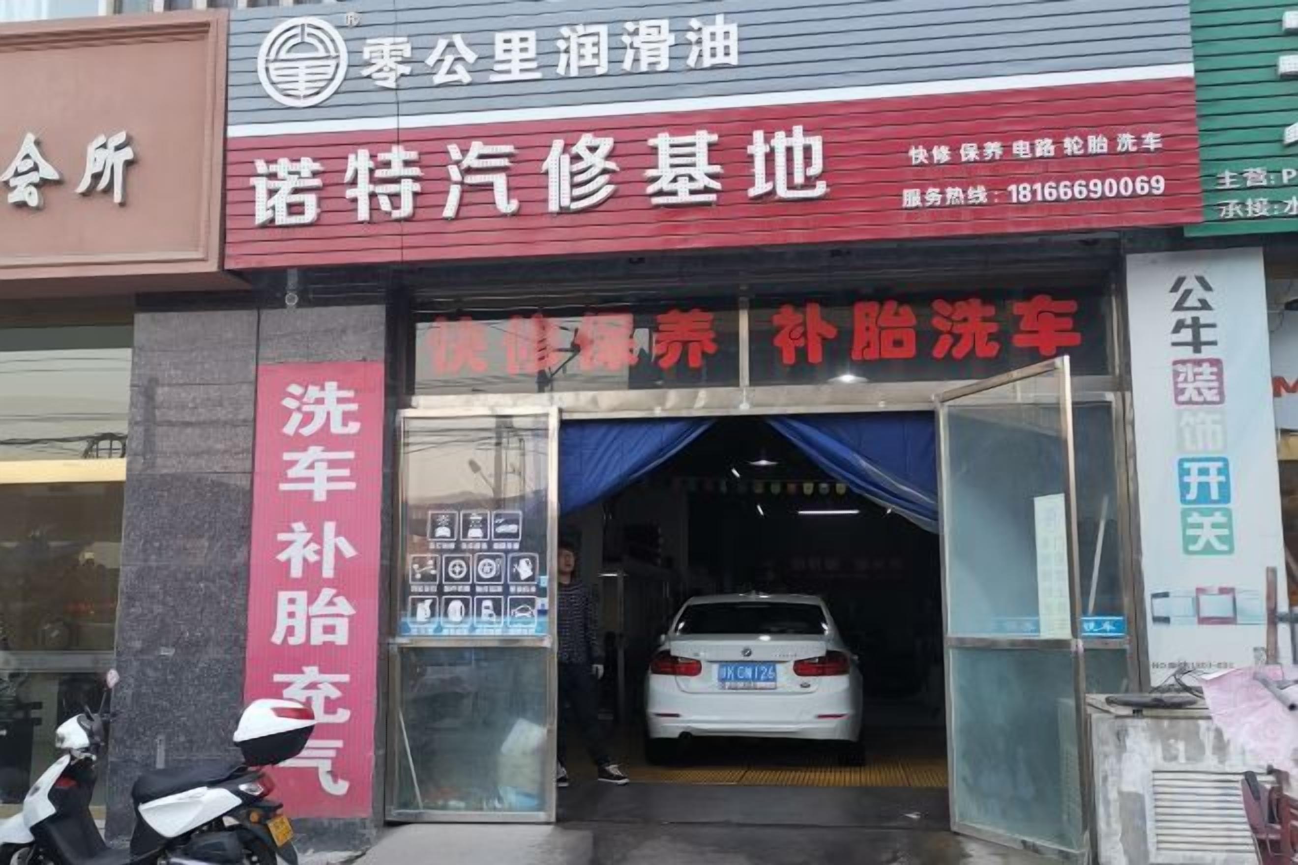 Un lavadero de coches con un letrero rojo y blanco, un coche blanco en su interior y una moto estacionada afuera.
