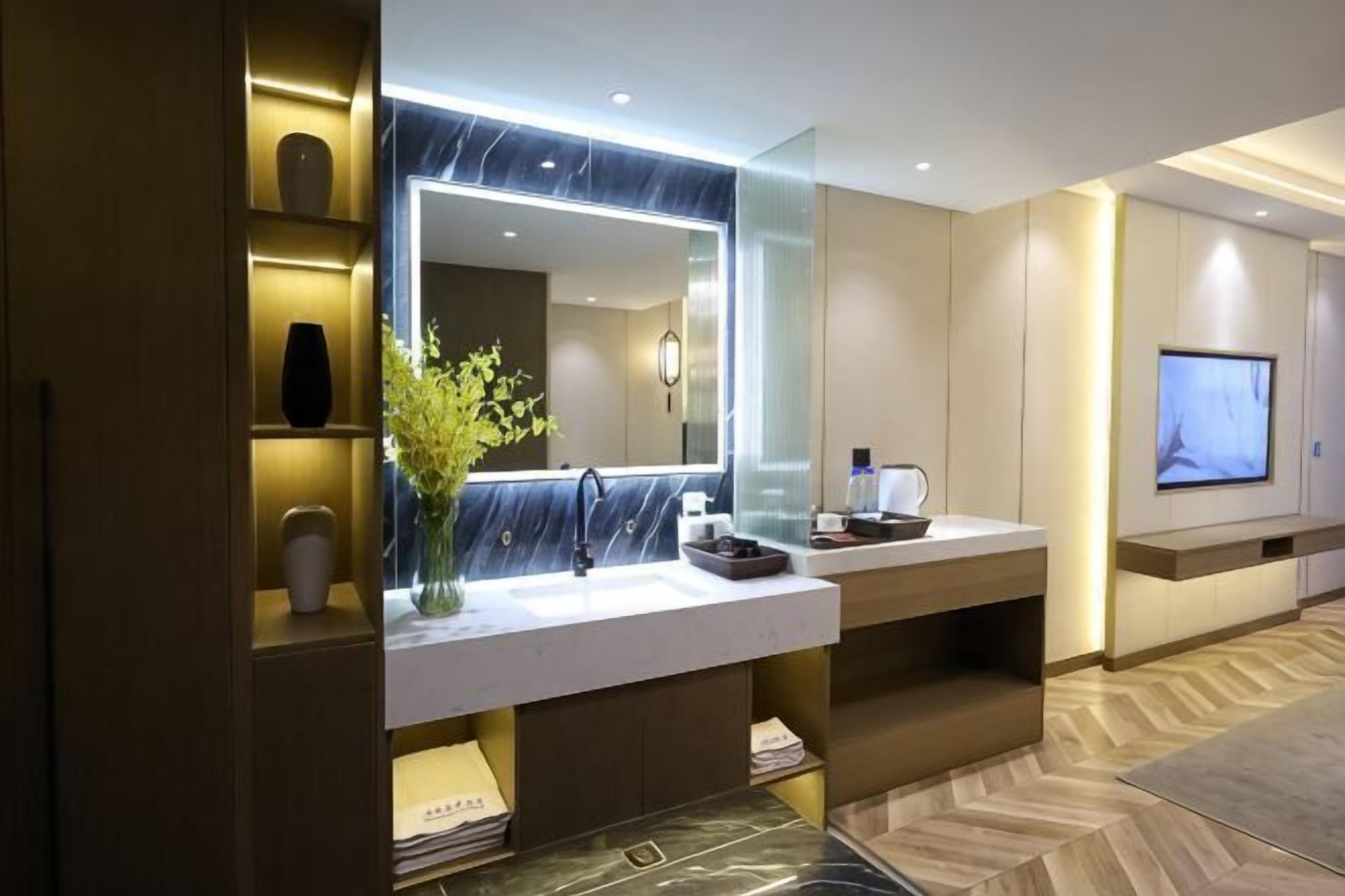 A modern bathroom with a large mirror, marble countertop, and a flat-screen TV mounted on the wall.. Shiji Jiamei Hotel