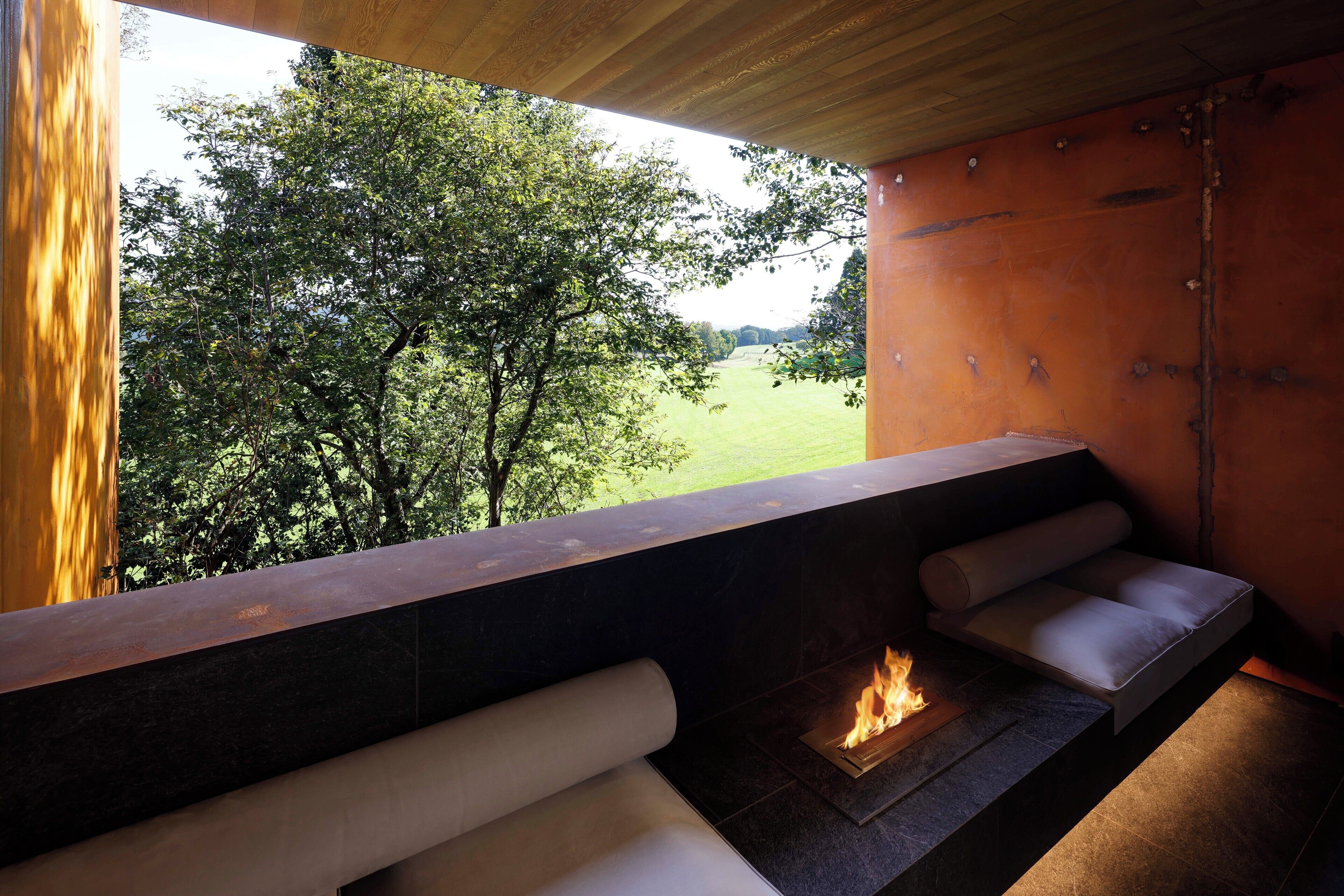 A modern outdoor seating area with a built-in fireplace, a view of a green landscape, and a wooden ceiling.
