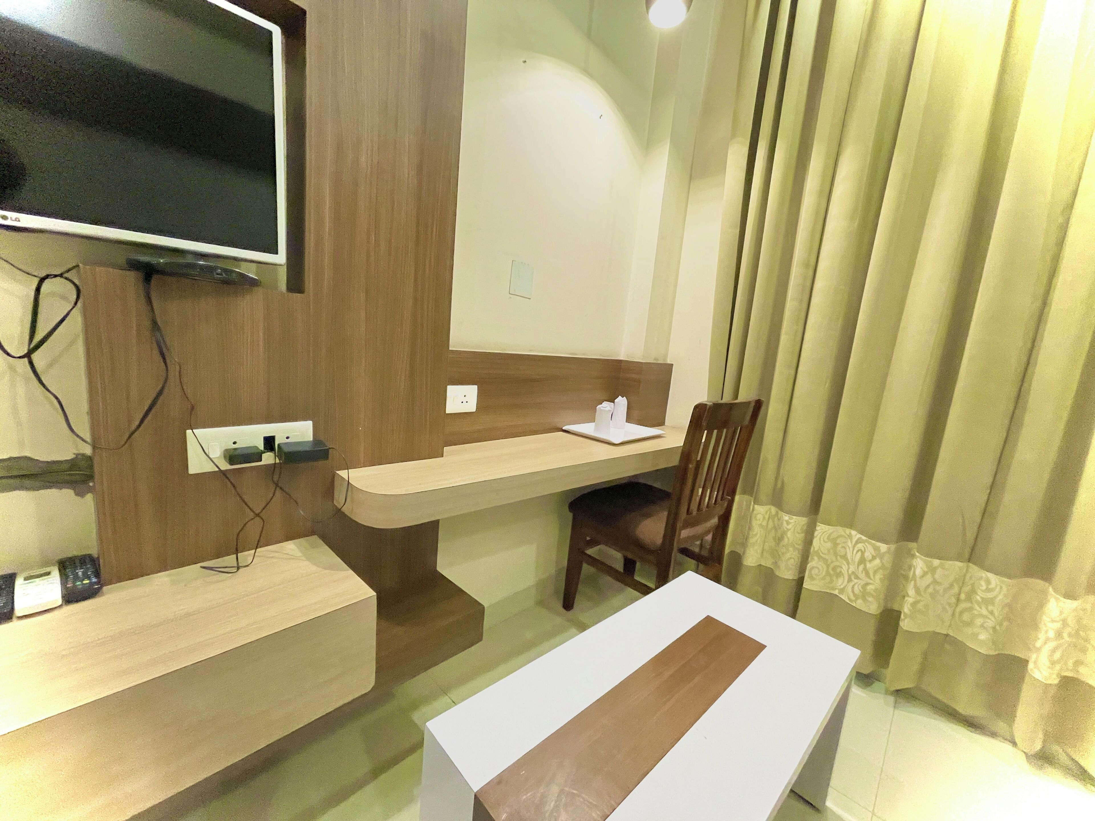 A hotel room with a wooden desk, a television mounted on the wall, a chair, and a bed.