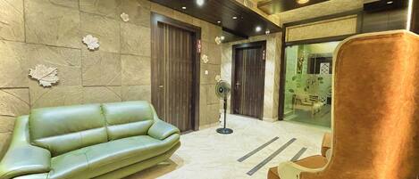A lobby with a green leather sofa, a brown armchair, and a fan.