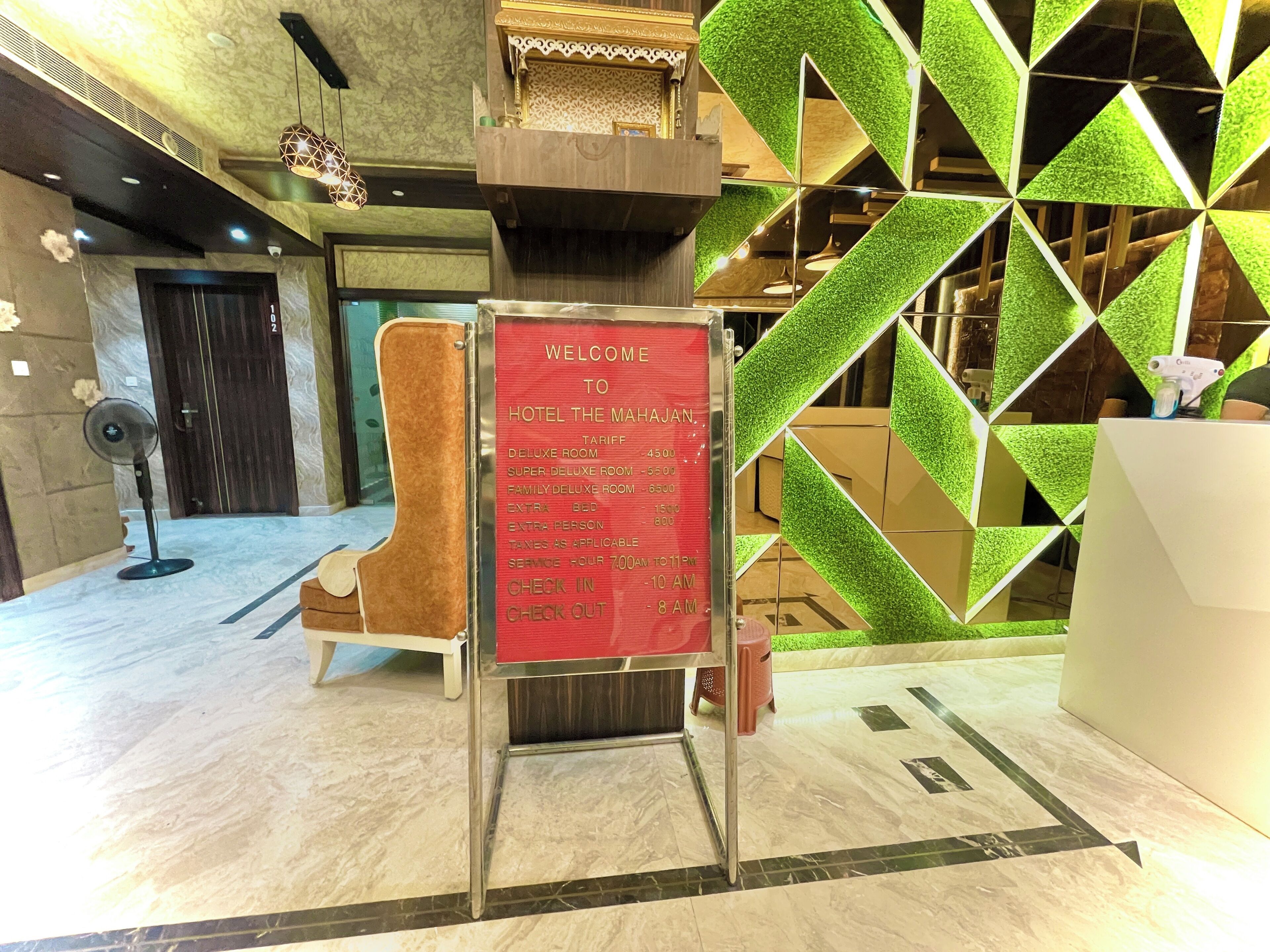 Zona de recepción de hotel con mostrador de registro, un ventilador y un panel decorativo con patrones geométricos.
