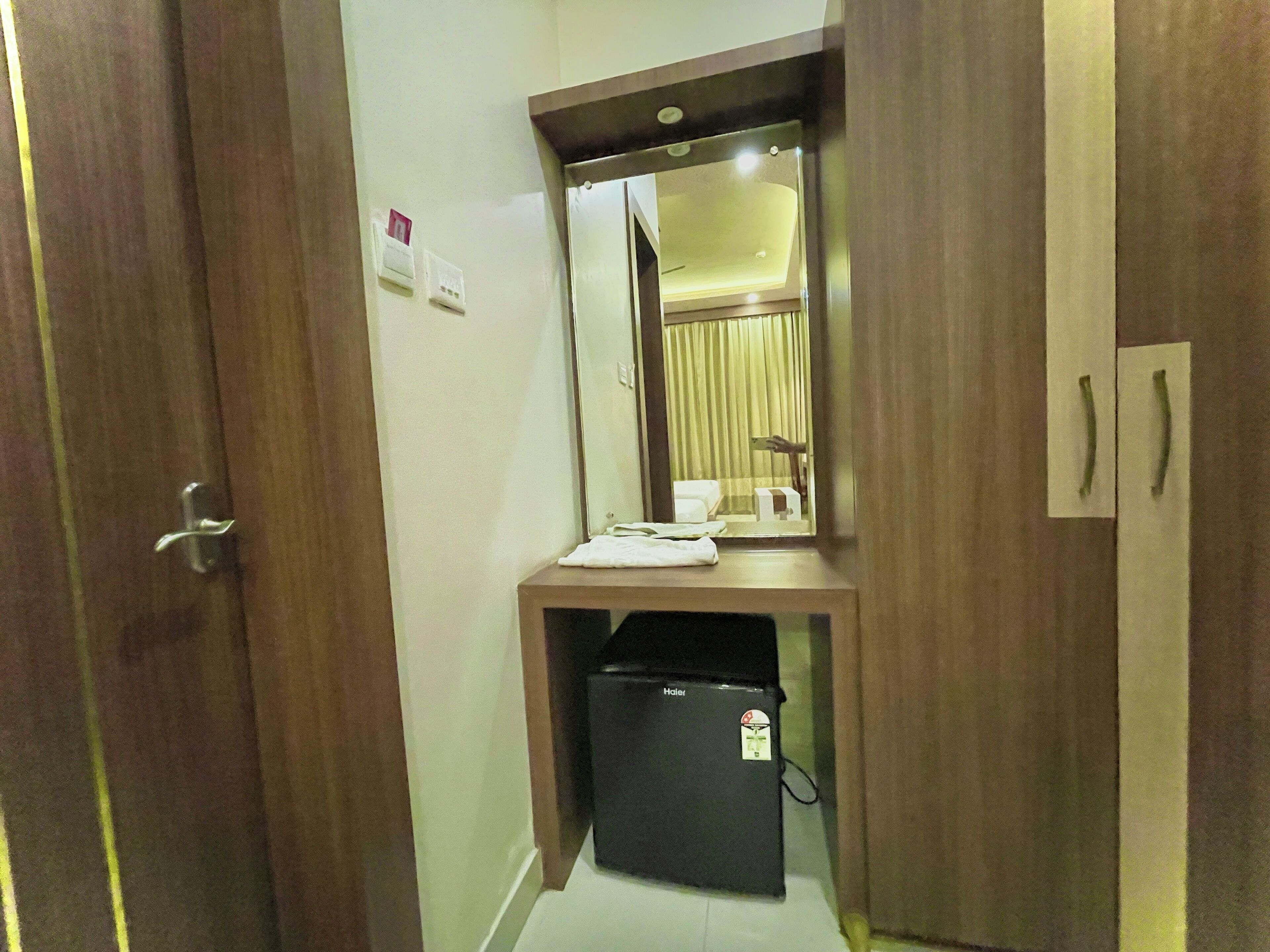 Ein Hotelzimmer mit Holztür, Spiegel über einem kleinen Schreibtisch, schwarzem Papierkorb und weißem Handtuch auf dem Schreibtisch.