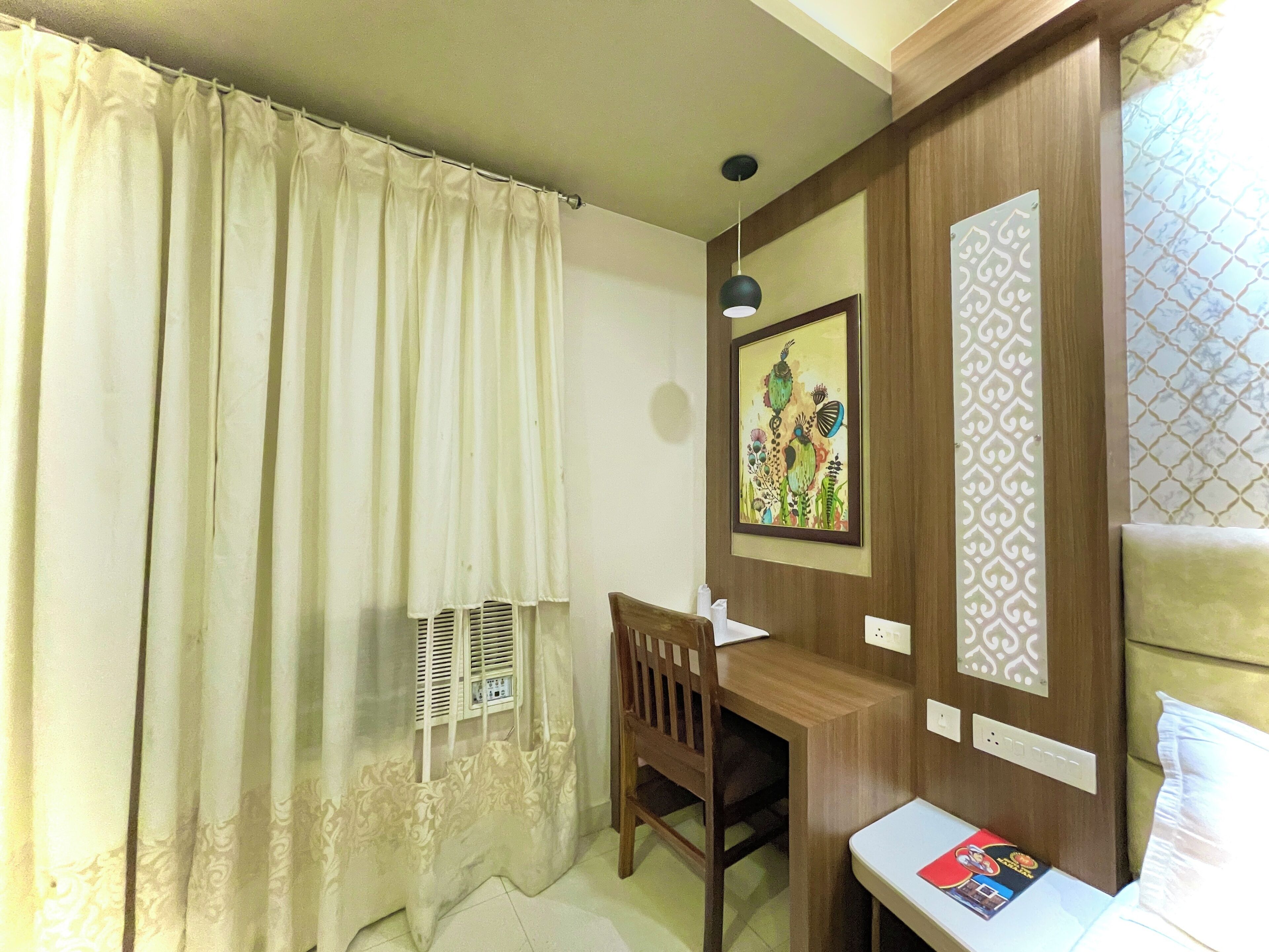 Una habitación de hotel con un escritorio de madera, una silla, una cama con cabecera verde, una ventana con cortina estampada y una obra de arte colgada en la pared.