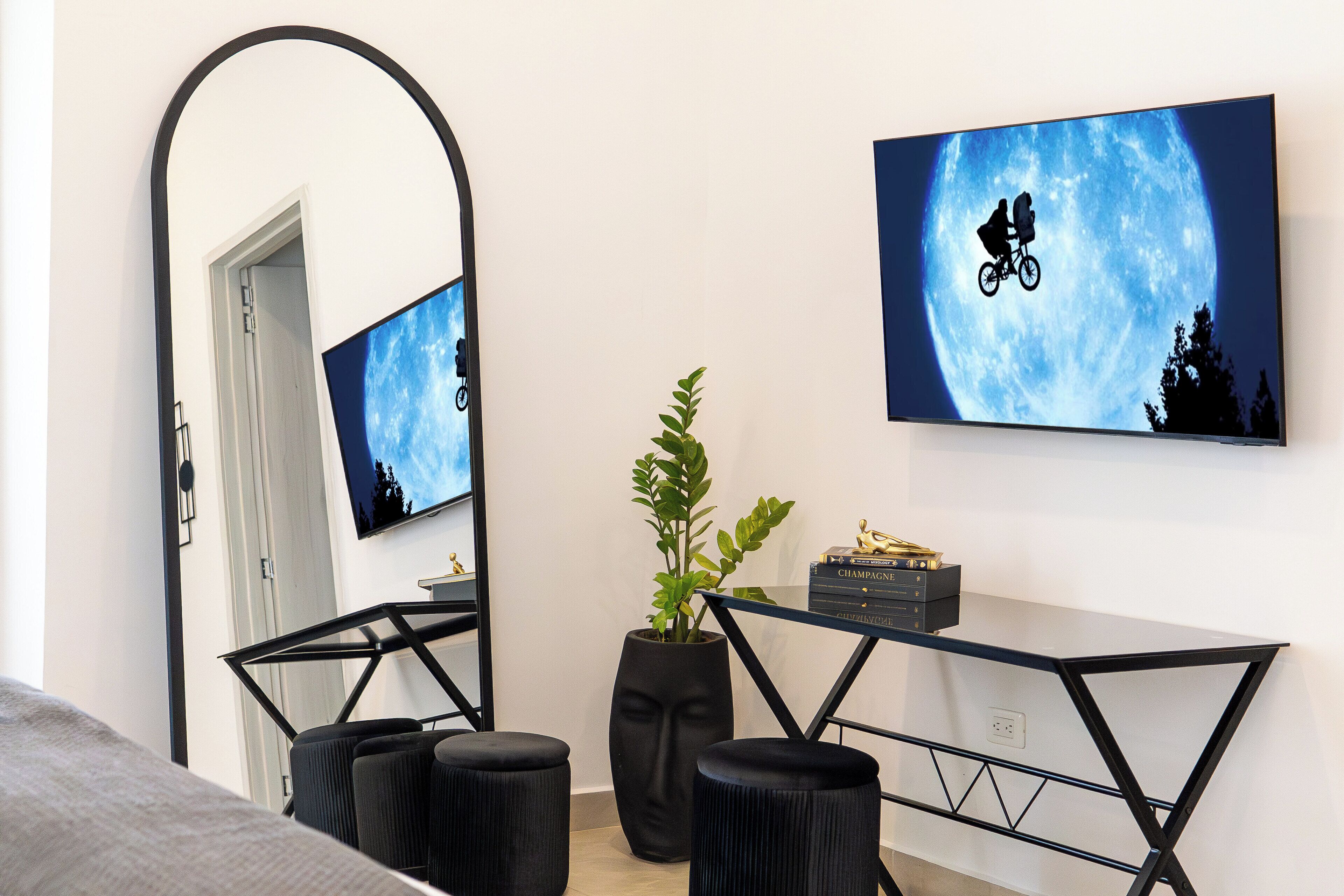 Une chambre moderne avec un grand miroir, un téléviseur affichant une silhouette de lune et de vélo, une plante en pot et une petite table sur laquelle se trouvent un livre et des objets décoratifs.
