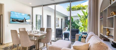 A modern living room with a dining area, large sliding glass doors leading to a balcony, and a television mounted on the wall.