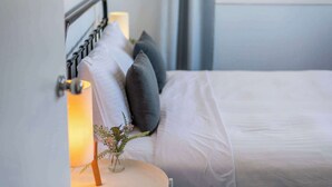A neatly made bed with white linens and a small bedside table with a lamp and a vase of flowers. - Blue House on Pippi - Oceanstays - Pet Friendly (Yamba)