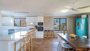 A modern kitchen with white cabinets, a wooden island with bar stools, a teal cabinet, and a large window. - Blue House on Pippi - Oceanstays - Pet Friendly (Yamba)