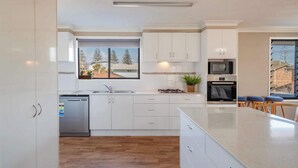 A modern kitchen with white cabinets, a stainless steel dishwasher, a microwave, and a large island with bar stools. - Blue House on Pippi - Oceanstays - Pet Friendly (Yamba)